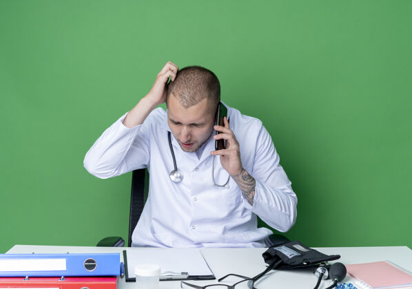 眼睛困惑的年轻男医生穿着医用长袍 手持听诊器坐在办公桌旁 手放在头上讲电话 眼睛闭在绿色的墙上男性穿头