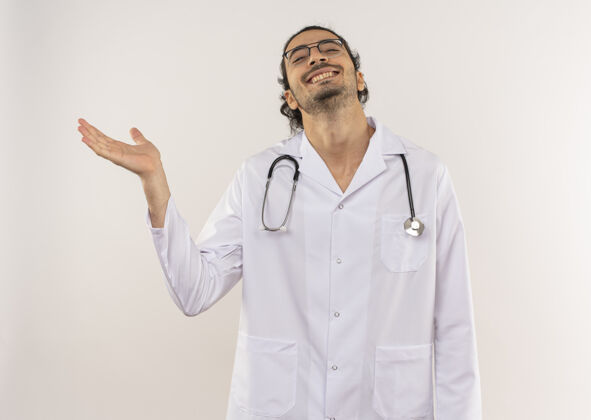 长袍面带微笑的年轻男医生戴着眼镜 身穿白色长袍 手持听诊器 将手指放在隔离的白色墙壁上 留有复印空间点穿壁板