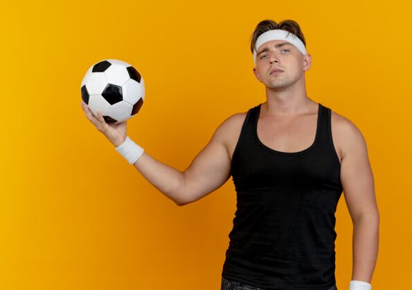 年轻自信的年轻帅气的运动型男人戴着头带和手环拿着足球看起来孤立在橙色头带足球橙色
