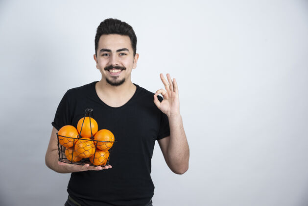 成人年轻人拿着装满橘子水果的金属篮子做着“ok”的手势人食物模型