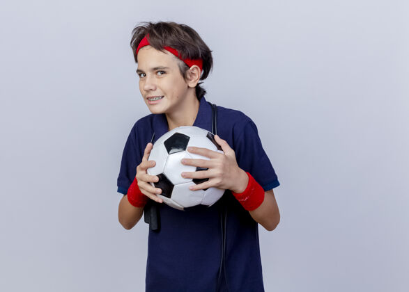 年轻狡猾的年轻英俊的运动男孩戴着头带和带牙套的腕带 脖子上系着跳绳 手里拿着足球 看着隔离在白色背景上的相机 还有复制空间持有男孩绳子