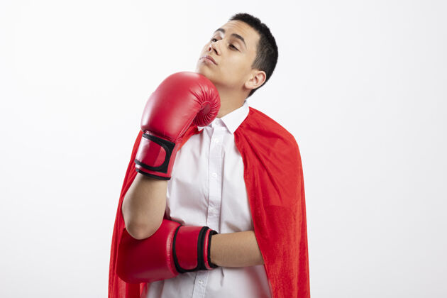 思想体贴的年轻超级英雄男孩 穿着红色斗篷 戴着盒子手套 看着侧面 手放在下巴上 白色背景上 留有复制空间手穿年轻