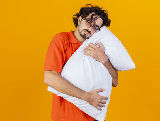 男人疲惫的年轻白种人 戴着眼镜 抱着枕头 把头放在上面 闭着眼睛 隔离在橙色背景上 留着复印空间复制拥抱年轻