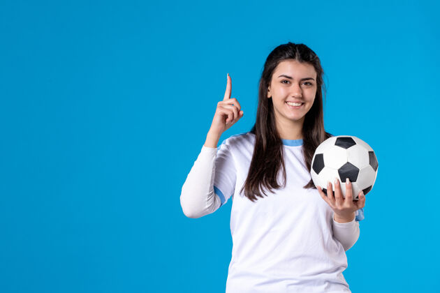 成年人前视图：蓝色墙上有足球的年轻女性球员前面视图