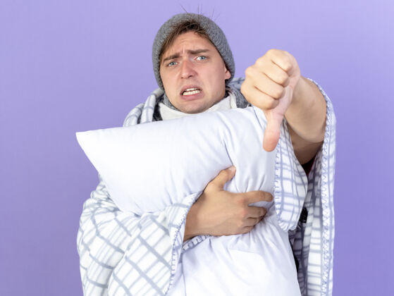 年轻皱眉的年轻英俊的金发病男子戴着冬天的帽子和围巾包裹在格子布抱枕看着相机显示拇指向下孤立的紫色背景显示疾病枕头