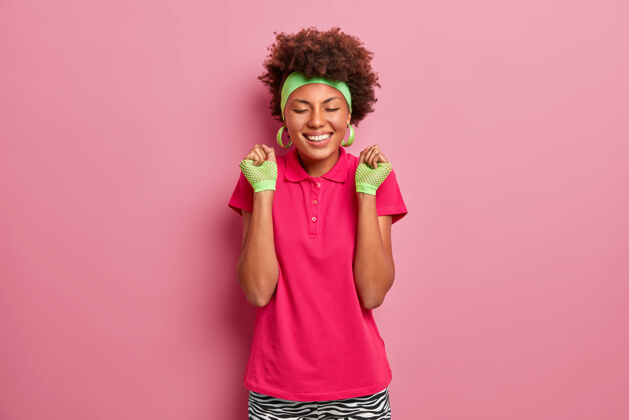 快乐快乐的情绪和感觉微笑的黑人女孩穿着粉色t恤 戴着运动手套和头带 高兴地握紧拳头 感受胜利的滋味 庆祝比赛胜利赢家高兴庆祝