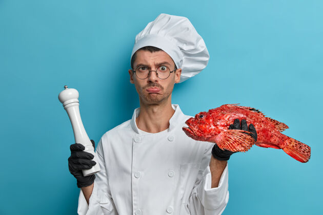 人不高兴累了的厨师拿着胡椒磨和新鲜的红鲈鱼 准备在烤架上用香料烤鱼 穿上制服 用盐调味悲伤食物鱼