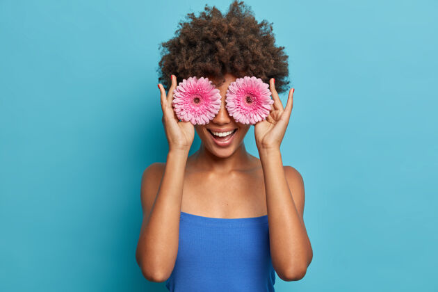 封面快乐的年轻美国黑人女孩的画像用玫瑰色的非洲菊遮住眼睛 玩得很开心 拿着最喜欢的花 有牙齿的微笑 穿着蓝色上衣 享受业余时间夏天芳香满意