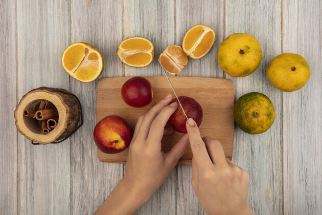 新鲜俯视图中 女性的手用刀在木制厨房板上切桃子 橘子被隔离在灰色的木墙上厨房橘子有机