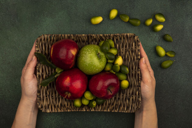 柳条顶视图中的女性手拿着柳条托盘的新鲜苹果与金冈隔离在绿色的墙上苹果托盘新鲜