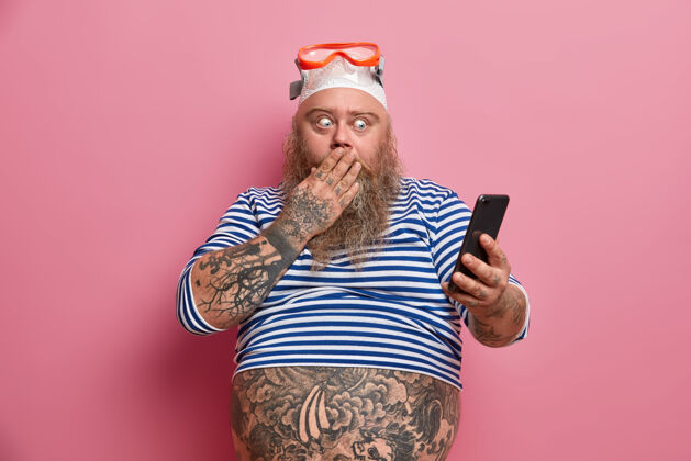 腹部身材魁梧 满脸胡须的男人捂着嘴 盯着智能手机 读着一些惊人的东西 穿着水手衬衫 戴着泳镜 肚子上有纹身夏天潜水员男人