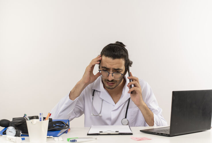 工具悲伤的年轻男医生戴着医用眼镜穿着医用长袍带着听诊器听诊器笔记本电脑男性