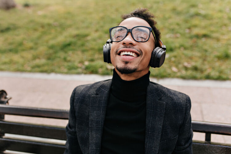放松笑眯眯的非洲男人在绿草地上的木凳上摆姿势戴着眼镜的快乐黑人闭着眼睛微笑着听音乐城市微笑享受