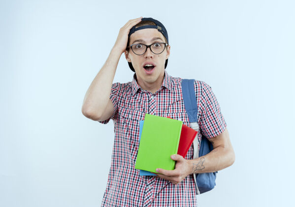 帽子惊讶的年轻学生男孩戴着背包 眼镜和帽子拿着书 把手放在白色的头上惊喜包背景