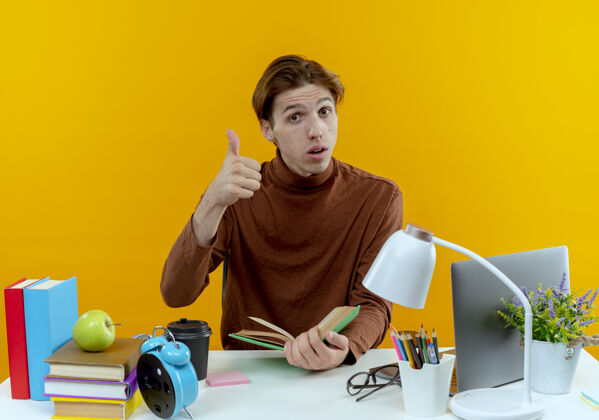 工具想着那个坐在书桌旁拿着学习工具拿着书的年轻学生 他的大拇指在黄色上书桌背景学校