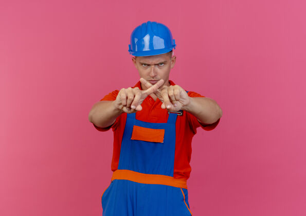 手势严格的年轻男性建筑工人穿着制服 戴着安全帽 在粉色上显示出“不”的手势安全严格头盔