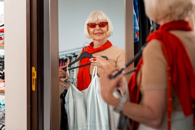 老年人永远年轻腰向上的老年时尚女子戴着红色太阳镜 看着购物镜 手里拿着夏装 一边欣赏她的倒影外观时尚商业