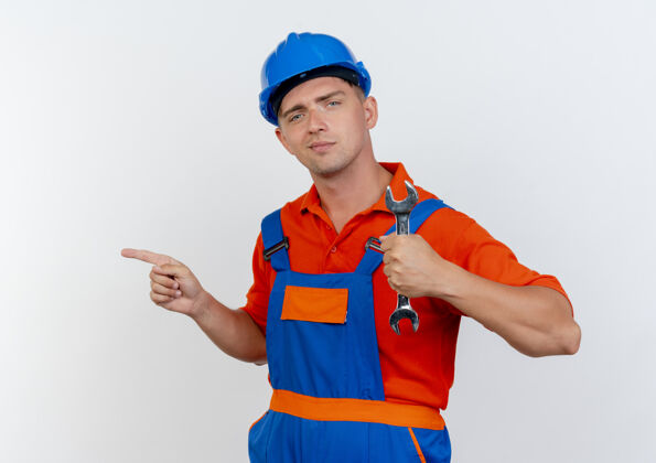 安全高兴的是年轻的男建筑工人穿着制服 戴着安全帽 手持扳手 指着旁边的白色制服壁板请