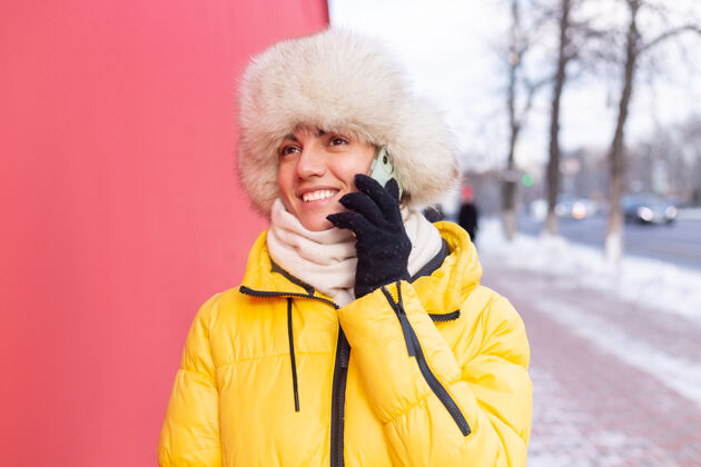 乐趣在一个冬天阳光明媚的日子里 一个穿着暖和衣服的红墙背景上的快乐的年轻女子在雪城的人行道上微笑着打电话肖像女孩人