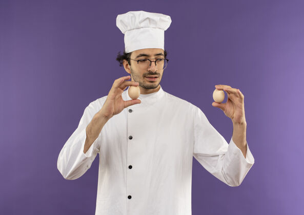 年轻想着年轻的男厨师穿着厨师制服 戴着眼镜拿着紫色的鸡蛋看着眼镜厨师持有
