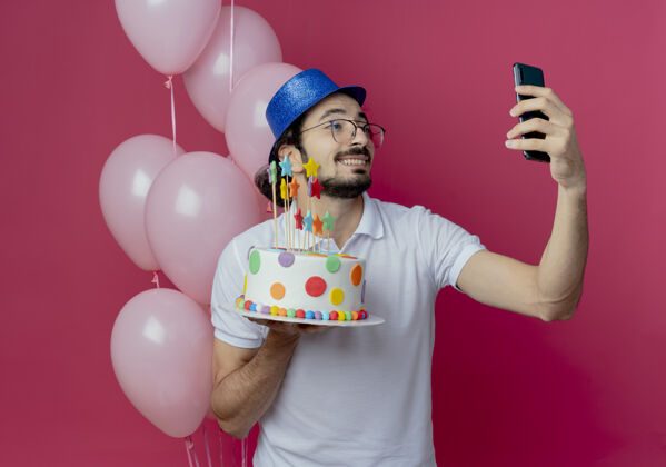 抱着笑容可掬的帅哥戴着眼镜和蓝色帽子站在气球前拿着蛋糕 在粉色上自拍站着帅哥微笑