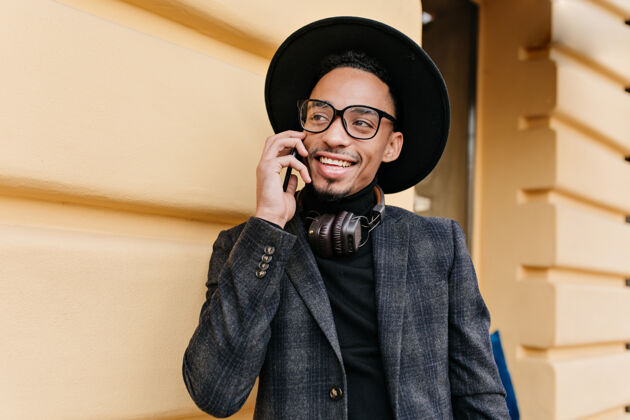 时尚快乐的非洲男人 大眼睛 一边叫着朋友 一边笑着一张穿着时髦的灰色衣服 拿着电话站在黄色墙壁边的照片手机帅气电话