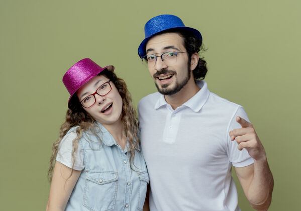 橄榄色微笑的年轻夫妇戴着粉色和蓝色的帽子 男人在橄榄绿上向你展示孤立的姿势穿着绿色帽子
