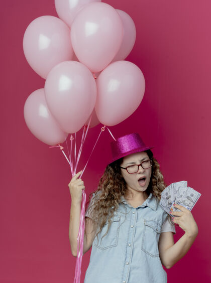 不愉快未出租的年轻女孩戴着眼镜和粉红色的帽子拿着气球和现金孤立在粉红色眼镜穿着帽子