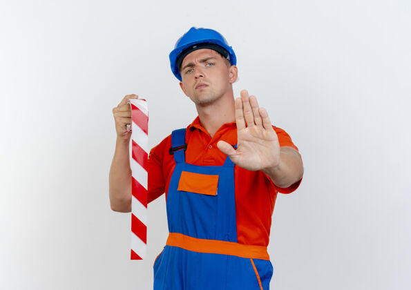 严格严格的年轻男性建筑工人穿着制服 戴着安全帽 拿着胶带 在白色地板上做停车手势胶带手持制服