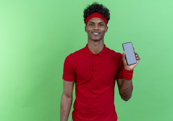 腕带高兴的年轻黑人美国运动男子戴着头带和手环举行电话隔离在绿色的墙壁上穿运动电话