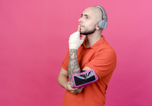 耳机看着一边思考的年轻人 手腕缠着绷带 戴着带电话臂带的耳机 手放在粉红色墙上的下巴上下巴运动墙