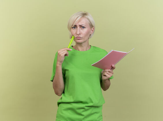 感情困惑的中年金发女人拿着便笺簿 用橄榄绿的墙上孤立的笔看着前面触摸的脸颊女人脸姿势