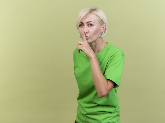 表情自信的中年金发女人站在侧视图中看着前面做保持沉默的手势隔离在橄榄绿的墙上衣服人自信