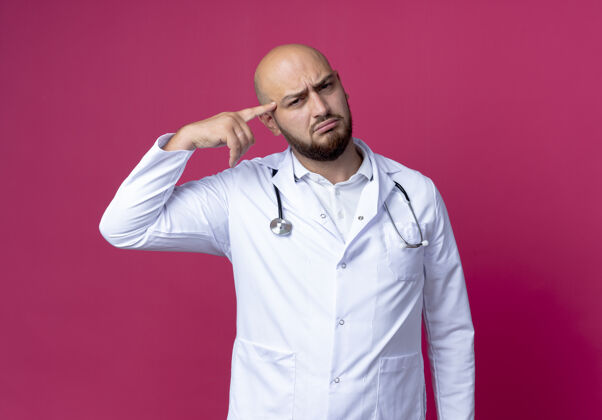 墙困惑的年轻秃头男医生穿着医用长袍和听诊器把手指放在额上隔离在粉红色的墙上年轻额头手指