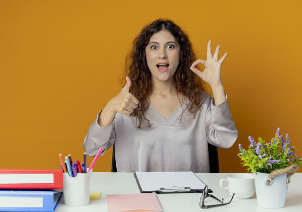 女人快乐的年轻漂亮的女上班族坐在办公桌旁 手里拿着办公工具 在橘色的墙上 她竖起大拇指做着一个很好的手势墙拇指手势