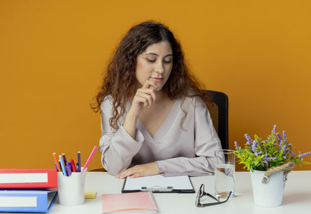 墙看着书桌心想：年轻漂亮的女上班族坐在书桌旁 手里拿着办公工具 把手指放在橘黄色的墙上手势年轻漂亮