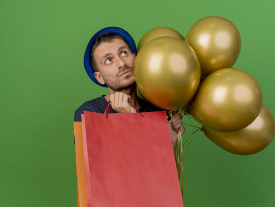 焦虑戴着蓝色派对帽的焦急帅哥拿着氦气球和纸购物袋 孤零零地看着绿色的墙上衣服感情气球