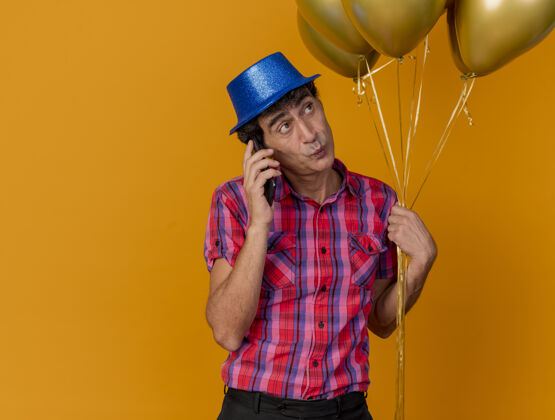 黄金印象深刻的中年党人戴着党的帽子拿着气球讲电话看着橙色墙上孤立的一面气球拿着人