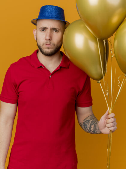 抱着愁眉不展的年轻帅哥戴着派对帽拿着气球看着孤立在橙色墙上的前面表情聚会人