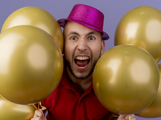 帽子特写镜头：愤怒的年轻帅哥戴着派对帽 看着前面拿着孤立在紫色墙上的气球聚会黄金男人
