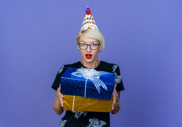生日给人印象深刻的年轻金发派对女郎戴着眼镜 戴着生日帽 拿着礼盒 看着前面孤立的紫色墙壁表情金发女人