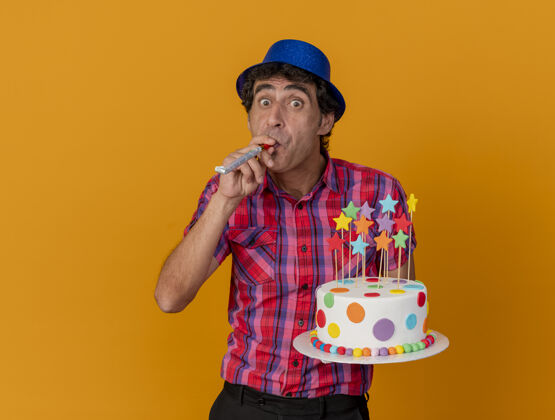 穿印象深刻的中年党人戴着党的帽子拿着生日蛋糕吹党吹风机看着前面孤立的橙色墙上人蛋糕吹