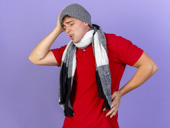帽子疼痛的年轻英俊的金发病夫戴着冬天的帽子和围巾 头和腰被隔离在紫色的墙上年轻衣服金发
