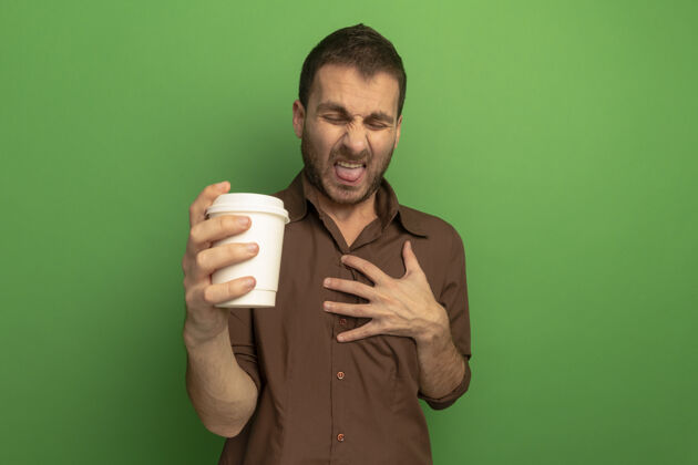 杯子恼怒的年轻人拿着塑料咖啡杯 手放在胸前 闭着眼睛 露出舌头 隔离在绿色的墙上拿着人人