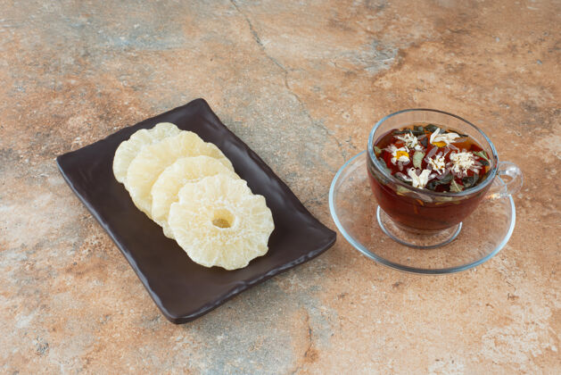 芳香的一个黑色的盘子里装满了健康的菠萝干和一杯茶干的杯子水果