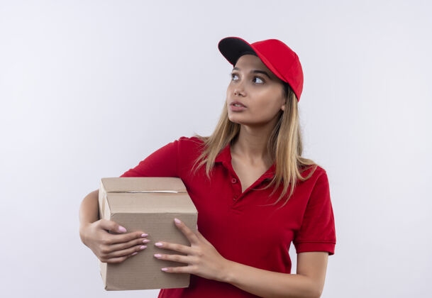 送货看着身边想着的年轻送货女孩 穿着红色制服 拿着帽子的盒子孤立在白色的墙上年轻穿壁板