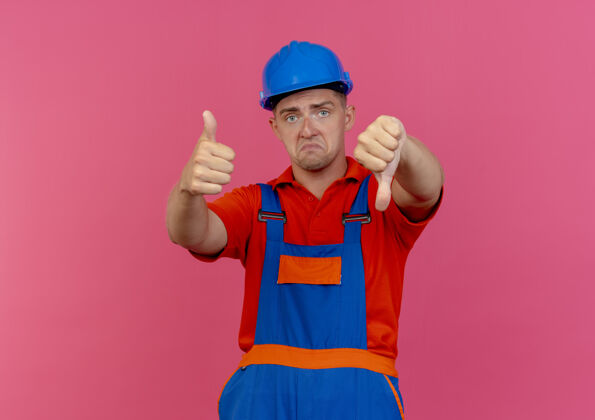 拇指悲伤的年轻男性建筑工人穿着制服和安全帽拇指上下粉红色安全向下头盔