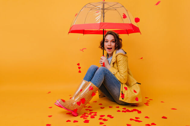 魅力穿着黄色胶鞋的情绪化的年轻女子坐在铺着纸心的地板上灵感四射的卷发女孩拿着可爱的雨伞摆姿势的室内照片年轻伞雨