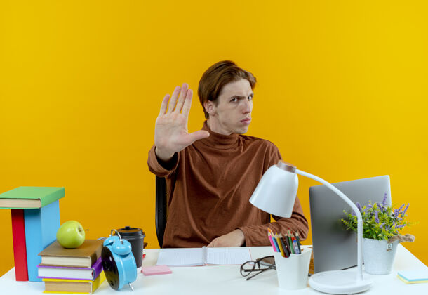工具严格的年轻学生男孩坐在书桌旁 手里拿着学习工具 在黄色上显示停止手势严格黄色手势