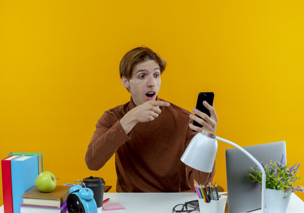 背景惊讶的年轻学生男孩坐在书桌旁 手里拿着学习工具 指着黄色的电话学生年轻男孩